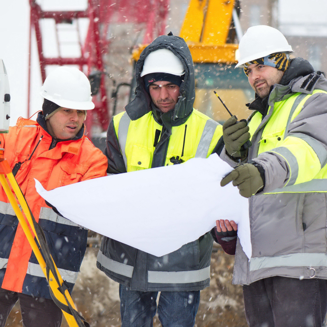 contractors wearing safety jackets, hardhats, and jackets looking at a large piece of paper together on a jobsite