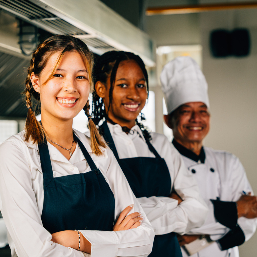 three restaurant kitchen workers in white button down shirts and black aprons lined up smilng in the kitchen with their arms folded over