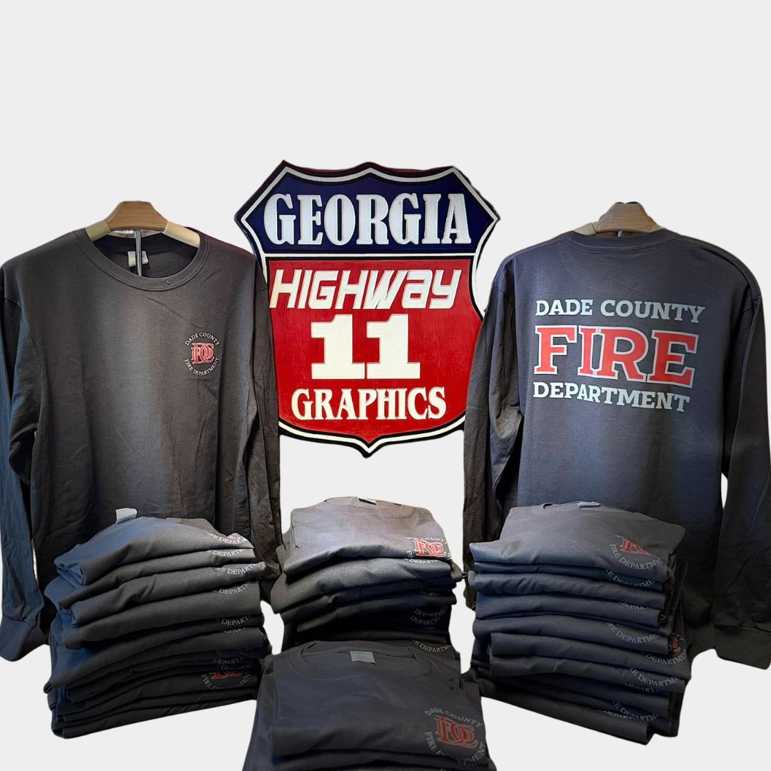 two long-sleeve shirts in between Highway 11 Graphics logo with a pile of folded shirts stacked below. All shirts are dark grey and have the logo for the Dade County Fire Department